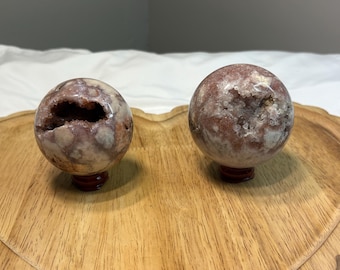 May include: Two spherical, polished stone geodes with a mix of purple, white, and brown hues. One geode has a large cavity. Both are displayed on small, dark wooden stands, set on a wooden tray.