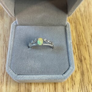 May include: A silver ring with a white opal gemstone and smaller clear gemstones set in a delicate crown design. The ring is displayed in a gray velvet ring box.