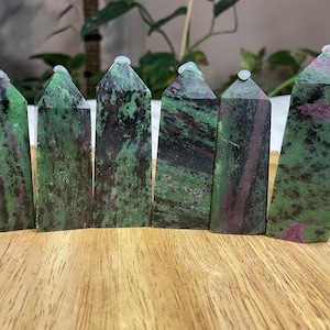 Puede incluir: Seis puntas de piedra preciosa verde, negra y rosa, también conocidas como zoisita, están dispuestas en una fila sobre una superficie de madera. Las puntas están pulidas y tienen un acabado liso y brillante.