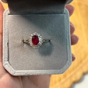 May include: A silver ring with a large oval ruby center stone, surrounded by smaller, clear stones. The ring is displayed in a gray velvet ring box, open to reveal the jewelry.