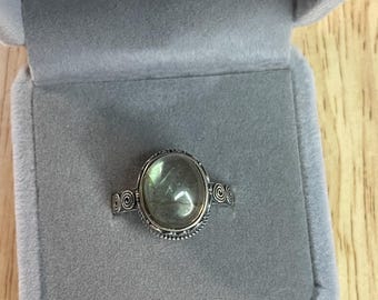 May include: A silver ring with a green gemstone set in a decorative bezel. The ring is in a gray velvet ring box.
