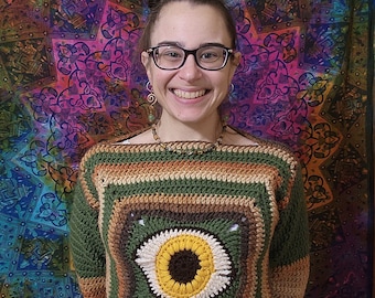 May include: A green, brown, and beige crocheted sweater with a large sunflower design in the center. The sweater has a square shape and is made with a chunky yarn.