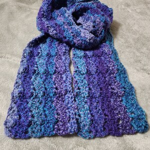 May include: A crocheted scarf in shades of purple and blue. The scarf is made with a textured stitch and has a fringed edge.