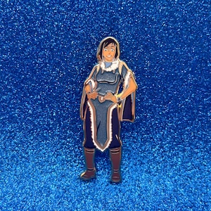 May include: Enamel pin featuring a character in a blue and brown outfit with a white fur-trimmed collar and cape. The character is standing with hands on hips, and brown boots. The pin is set against a sparkly blue background.