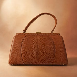 May include: A structured handbag in a rich tan colour featuring a textured lizard-skin pattern. The bag has a single arched top handle, a rectangular frame, and a small gold-toned metal clasp closure at the top centre of the main compartment.