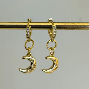 May include: Gold-coloured hoop earrings with a crescent moon charm. The hoops are adorned with small, clear stones. The crescent moons have a textured surface and are also embellished with small, clear stones. The earrings hang from a gold bar.