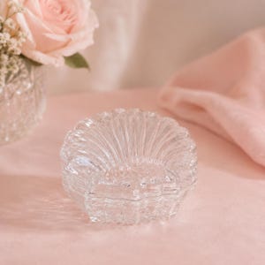 May include: A clear glass trinket dish shaped like a scallop shell with a ribbed texture and a scalloped edge. The dish features a hinged lid design and a transparent finish, reflecting light across its curved surface and detailed ridges.