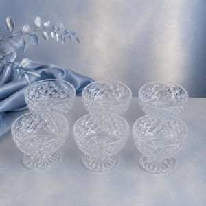 May include: A set of six clear glass dessert bowls featuring a diamond-cut pattern on the exterior and a starburst design on the base. Each bowl is supported by a short, thick pedestal stem, providing a stable foundation for serving various treats.