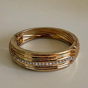 May include: Gold-coloured hinged bangle bracelet with a textured, ridged design. A row of small, clear crystals is set into the bracelet. The bracelet is a medium size and appears to be made of metal.