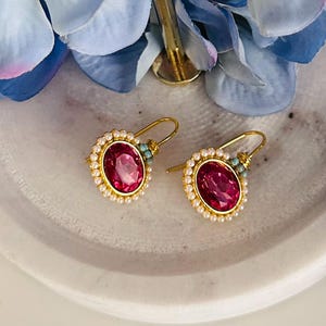 May include: Two gold-tone earrings featuring oval, ruby-red gemstones. Each earring is encircled by tiny, white pearls and embellished with turquoise beads. The earrings are presented on a white, marble-effect surface, with blue flower petals in the background.