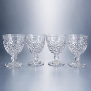 May include: A set of four clear crystal goblets featuring a deep diamond-cut pattern on the bowl, a short stem with a central spherical node, and a circular base with a starburst design etched into the underside of each glass vessel.