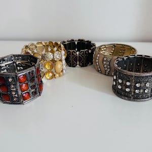 May include: Five stretch bracelets in different styles and colours. The bracelets include silver, gold, black, and red designs. Some are decorated with gemstones and other decorative elements, offering a variety of looks.