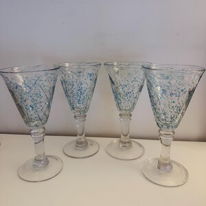 May include: A set of four clear glass goblets featuring a flared bowl with a speckled pattern of blue and teal flecks. Each goblet has a smooth, transparent stem and a circular base, providing a consistent design across all four drinking vessels.