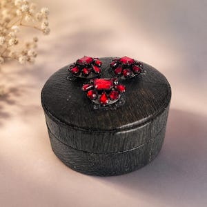 May include: A round, dark-stained wooden box with a textured grain finish. The lid is adorned with three clusters of faceted, deep red crystals arranged in a triangular pattern, each featuring a central rectangular stone surrounded by smaller gems.