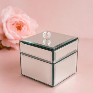 May include: A square mirrored jewellery box with bevelled glass edges and a clear, faceted crystal knob on the lid. The box features a reflective surface on all sides and the top, creating a clean, geometric appearance for storing small items.