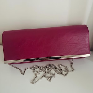 May include: A magenta clutch bag with a silver metal trim and a silver chain strap. The bag is rectangular and made of a textured material. The chain strap is draped on the surface. The design is simple and elegant.