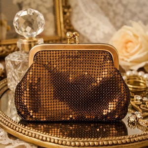May include: A vintage-style, bronze-colored metal mesh clutch with a gold-toned frame and clasp. The purse is open, revealing the interior. The clutch is on a gold-rimmed tray, with a perfume bottle and jewelry in the background.