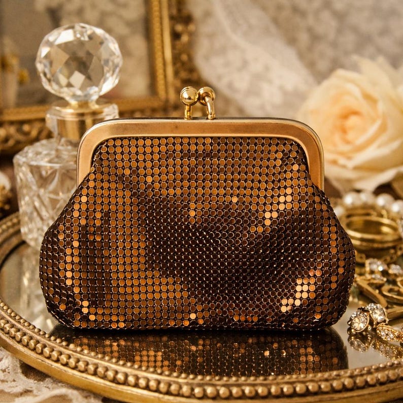May include: A vintage-style, bronze-colored metal mesh clutch with a gold-toned frame and clasp. The purse is open, revealing the interior. The clutch is on a gold-rimmed tray, with a perfume bottle and jewelry in the background.