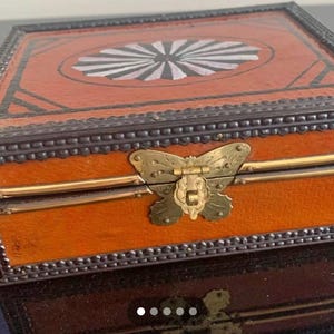 May include: A rectangular wooden box with a hinged lid. The box is stained orange with a decorative design on the lid. The lid features a white and black sunburst design. The box has a brass butterfly clasp and metal trim.
