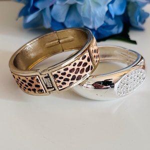 May include: Two hinged bangle bracelets. One features a gold-tone metal frame with a leopard print pattern. The other is silver-toned, with a cluster of clear crystals.
