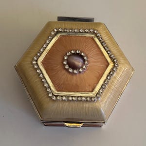 May include: A hexagonal compact mirror with a cream and peach enamel finish. The centre features a peach-coloured design with a central stone surrounded by small crystals. The edges are lined with additional crystals, and the compact has a silver metal frame.