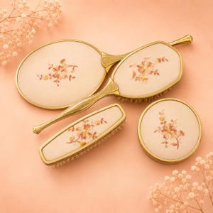 May include: A vanity set comprising an oval hand mirror, a hairbrush, a clothes brush and a round powder jar. Each item features a gold-tone metal frame and a cream-coloured fabric inlay embroidered with a delicate orange and yellow floral motif.