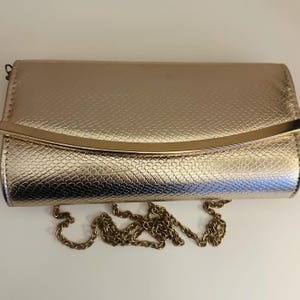 May include: A gold clutch bag with a textured, snakeskin-like pattern. The rectangular bag has a curved gold bar on the front and a gold chain strap. The clutch is closed, and the chain lies below it.