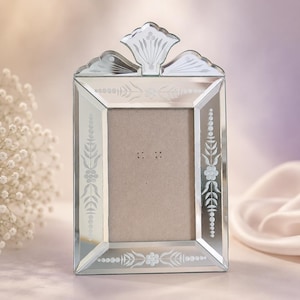 May include: A rectangular mirrored picture frame featuring bevelled edges and an ornate, arched top crest. The glass surface is decorated with delicate, etched floral and leaf patterns along the borders, surrounding a plain, light beige insert.