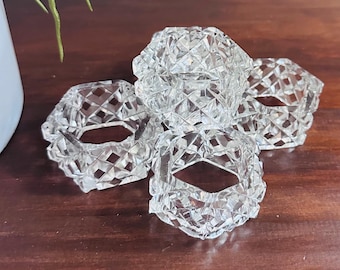 May include: Four clear, faceted crystal napkin rings. The rings are arranged on a dark brown wooden surface. The crystal rings have a geometric diamond pattern, reflecting light. The napkin rings are a decorative element for table settings.