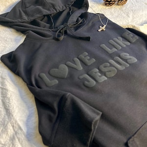 Può includere: Felpa con cappuccio nera con il testo "LOVE LIKE JESUS" in lettere in rilievo con un cuore al posto della "O".