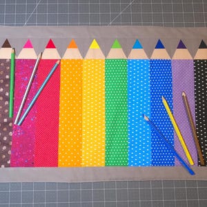 Pode incluir: Um colorido jogo americano com tema de lápis e borda cinza. O design apresenta uma fileira de lápis de várias cores, incluindo vermelho, laranja, amarelo, verde, azul e roxo. Vários lápis estão espalhados no tapete.