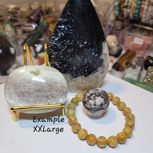 May include: A set of three crystal stones, including a black and white teardrop-shaped stone, a white and gray round stone, and a yellow and brown beaded bracelet. The stones are displayed on a gold stand. The text "Example XXLarge" is written on the white surface below the stones.