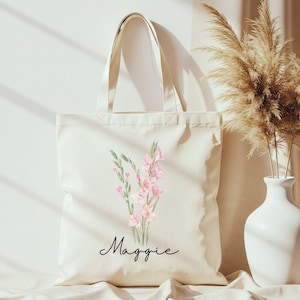 May include: Cream-colored tote bag featuring a watercolor floral design with pink and green accents. The name "Maggie" is written in a cursive font below the flowers. The bag has two cream-colored handles.
