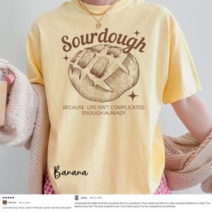 May include: Yellow t-shirt with a brown illustration of a sourdough loaf and the text "Sourdough BECAUSE LIFE ISN'T COMPLICATED ENOUGH ALREADY." The word "Banana" is written in cursive at the bottom.