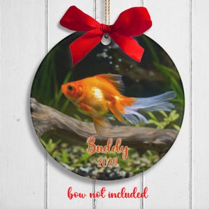 May include: Round Christmas ornament featuring a goldfish swimming in an aquarium. The ornament is decorated with the name "Buddy" and the year "2024".