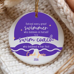 May include: A white ceramic ornament with a gold string for hanging. The ornament features a purple design with the text "Behind every great swimmer who believes in herself is a swim coach." It also includes the name "Coach Jessie" and the year "2026."