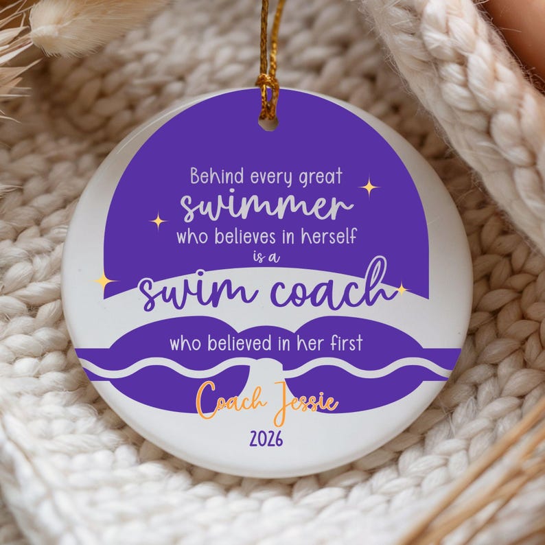 May include: A white ceramic ornament with a gold string for hanging. The ornament features a purple design with the text "Behind every great swimmer who believes in herself is a swim coach." It also includes the name "Coach Jessie" and the year "2026."