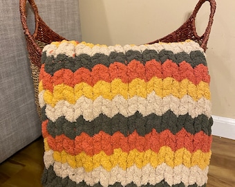 May include: A square cushion with a chevron pattern in cream, yellow, orange, and green. The cushion is made of a soft, chunky yarn. It is placed in front of a woven basket with handles.