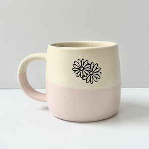 K&ouml;nnte beinhalten: Eine wei&szlig;e und rosafarbene Keramiktasse mit einem schwarzen G&auml;nsebl&uuml;mchen-Design. Die Tasse hat eine abgerundete Form und einen Henkel.