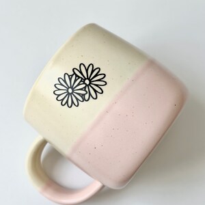 K&ouml;nnte beinhalten: Eine Keramiktasse mit zweifarbigem Glase, oben cremefarben und unten hellrosa. Die Tasse hat eine quadratische Form und ein schwarzes G&auml;nsebl&uuml;mchen-Design auf dem cremefarbenen Teil.