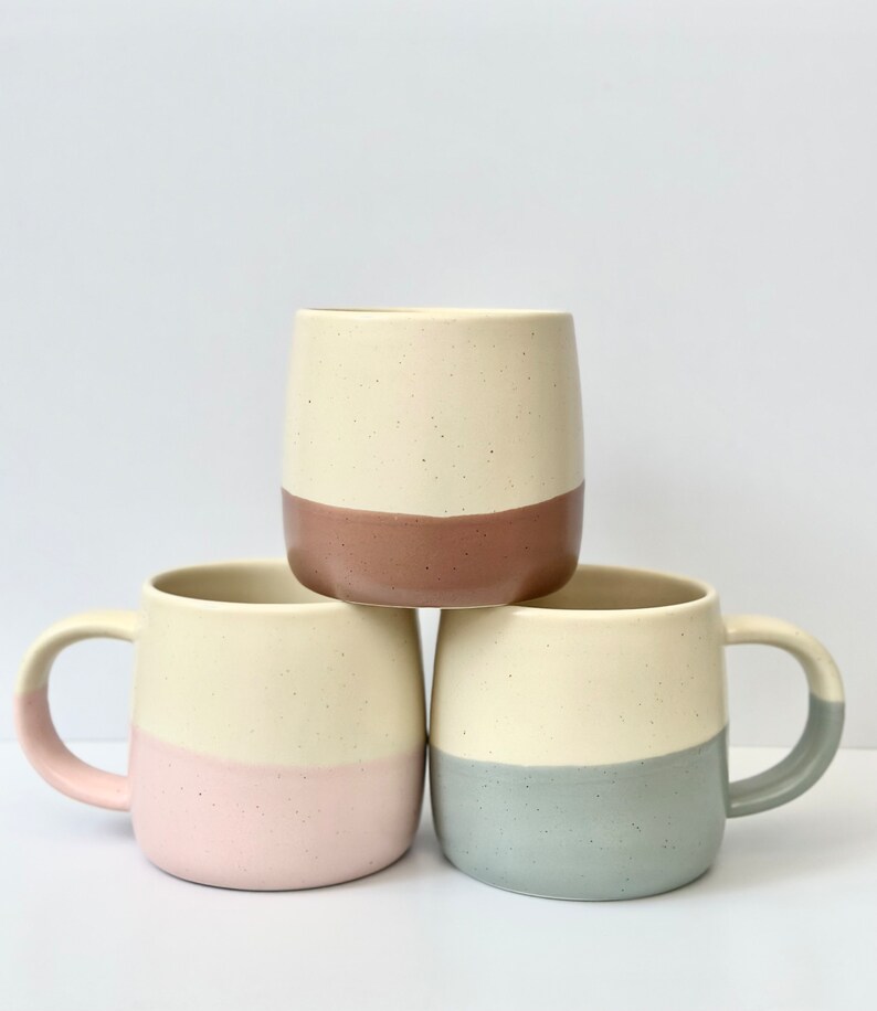 K&ouml;nnte beinhalten: Drei Keramikbecher mit gesprenkeltem wei&szlig;en &Uuml;berzug und einem farbigen Band am Boden. Die Becher sind &uuml;bereinander gestapelt. Der obere Becher hat ein braunes Band, der mittlere Becher ein rosa Band und der untere Becher ein hellblaues Band.