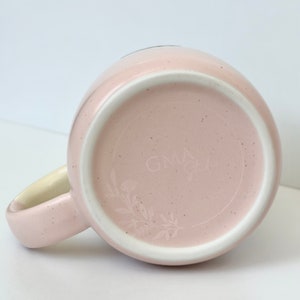 K&ouml;nnte beinhalten: Eine rosa Keramiktasse mit einem wei&szlig;en Henkel und einem floralen Design auf der Unterseite. Die Tasse hat den Text "GNLA" auf der Unterseite.