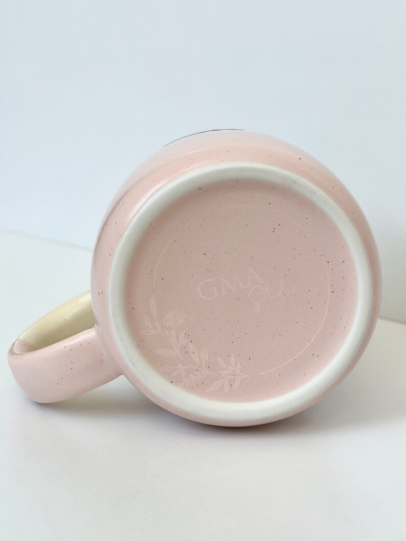 K&ouml;nnte beinhalten: Eine rosa Keramiktasse mit einem wei&szlig;en Henkel und einem floralen Design auf der Unterseite. Die Tasse hat den Text "GNLA" auf der Unterseite.