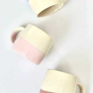 K&ouml;nnte beinhalten: Drei Keramikbecher mit zweifarbigen Designs. Die Becher sind wei&szlig; mit einem Farbband am Boden. Die Farben sind hellblau, rosa und braun.