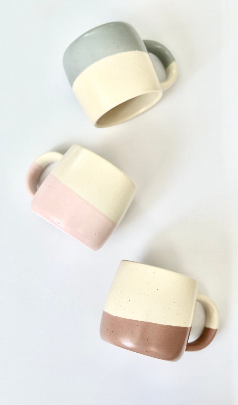 K&ouml;nnte beinhalten: Drei Keramikbecher mit zweifarbigen Designs. Die Becher sind wei&szlig; mit einem Farbband am Boden. Die Farben sind hellblau, rosa und braun.