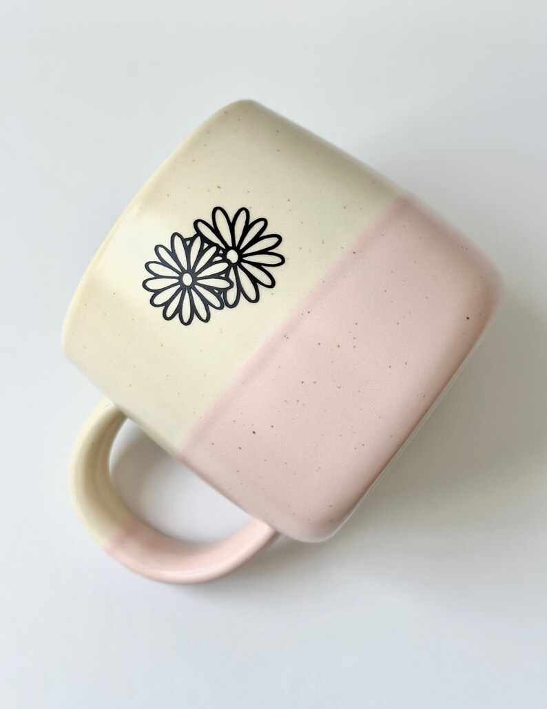 K&ouml;nnte beinhalten: Eine Keramiktasse mit zweifarbigem Glase, oben cremefarben und unten hellrosa. Die Tasse hat eine quadratische Form und ein schwarzes G&auml;nsebl&uuml;mchen-Design auf dem cremefarbenen Teil.