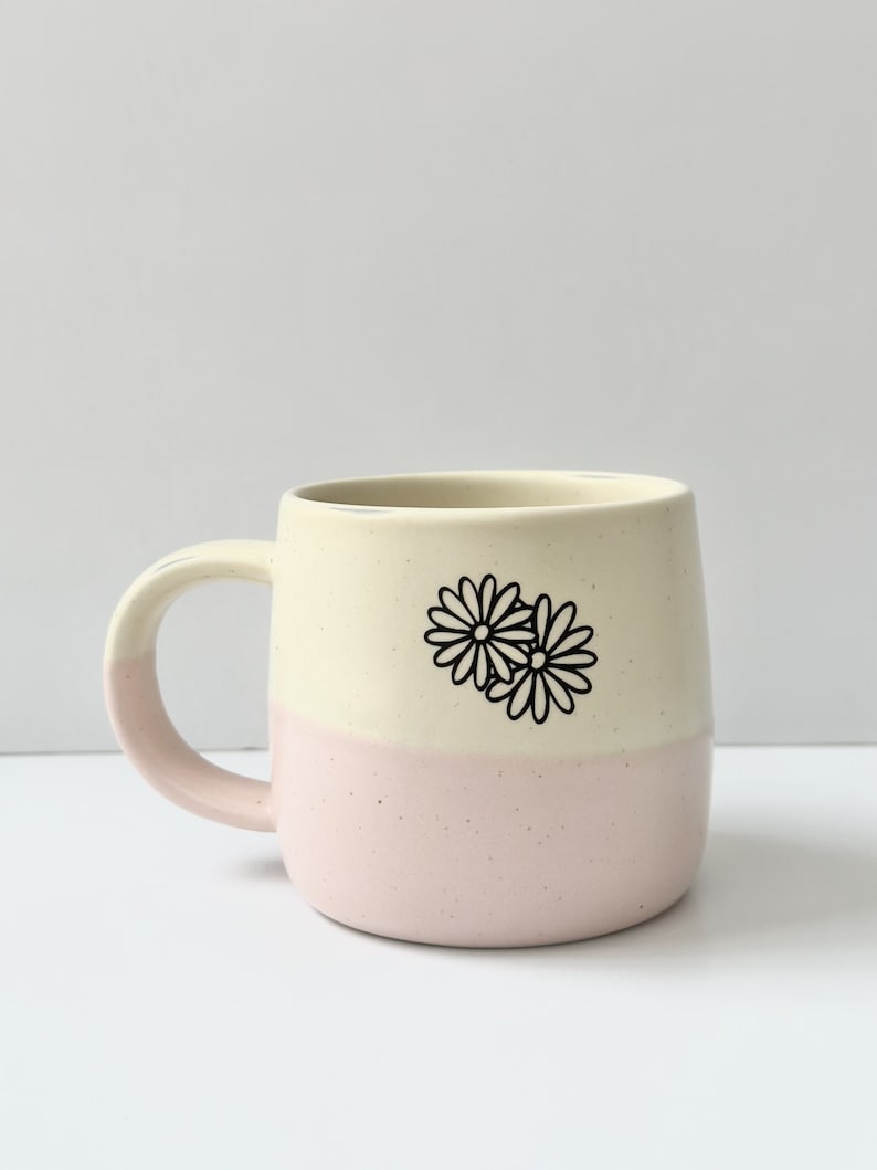K&ouml;nnte beinhalten: Eine wei&szlig;e und rosafarbene Keramiktasse mit einem schwarzen G&auml;nsebl&uuml;mchen-Design. Die Tasse hat eine abgerundete Form und einen Henkel.