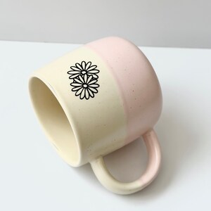 K&ouml;nnte beinhalten: Eine zweifarbige Keramiktasse mit cremefarbener und rosafarbener Glasur. Die Tasse hat einen Henkel und ein schwarzes G&auml;nsebl&uuml;mchen-Design.
