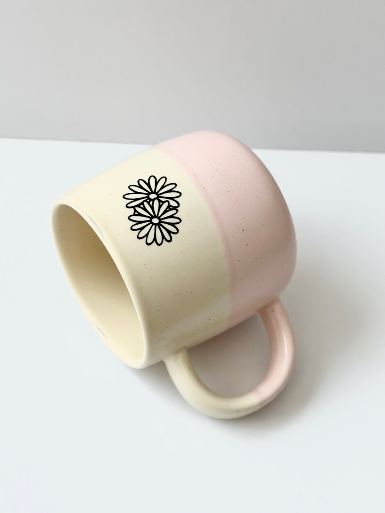 K&ouml;nnte beinhalten: Eine zweifarbige Keramiktasse mit cremefarbener und rosafarbener Glasur. Die Tasse hat einen Henkel und ein schwarzes G&auml;nsebl&uuml;mchen-Design.
