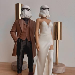 Puede incluir: Figuras de novios con cascos de Stormtrooper. El novio lleva un abrigo marrón y pantalones negros. La novia viste un vestido blanco con una abertura. Las figuras están sobre una superficie de madera.